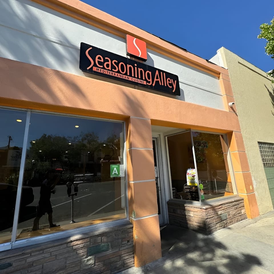 Exterior of Seasoning Alley Mediterranean Cuisine, with a black sign and orange trim under a blue sky.