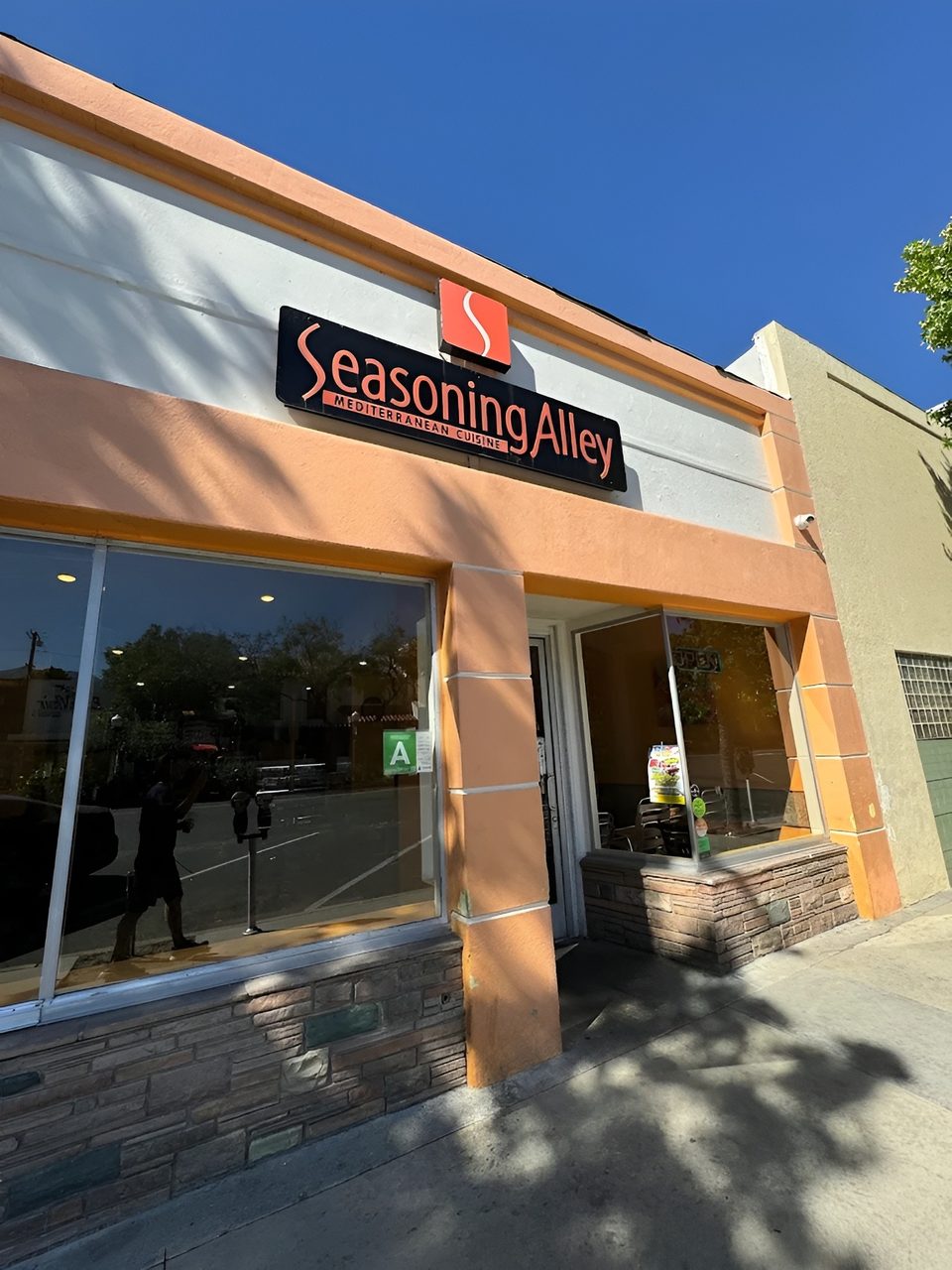 Exterior of Seasoning Alley Mediterranean Cuisine, with a black sign and orange trim under a blue sky.